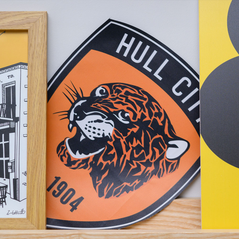 Orange and black Hull City emblem with a roaring tiger in profile, surrounded by a wooden photo frame and a yellow object.