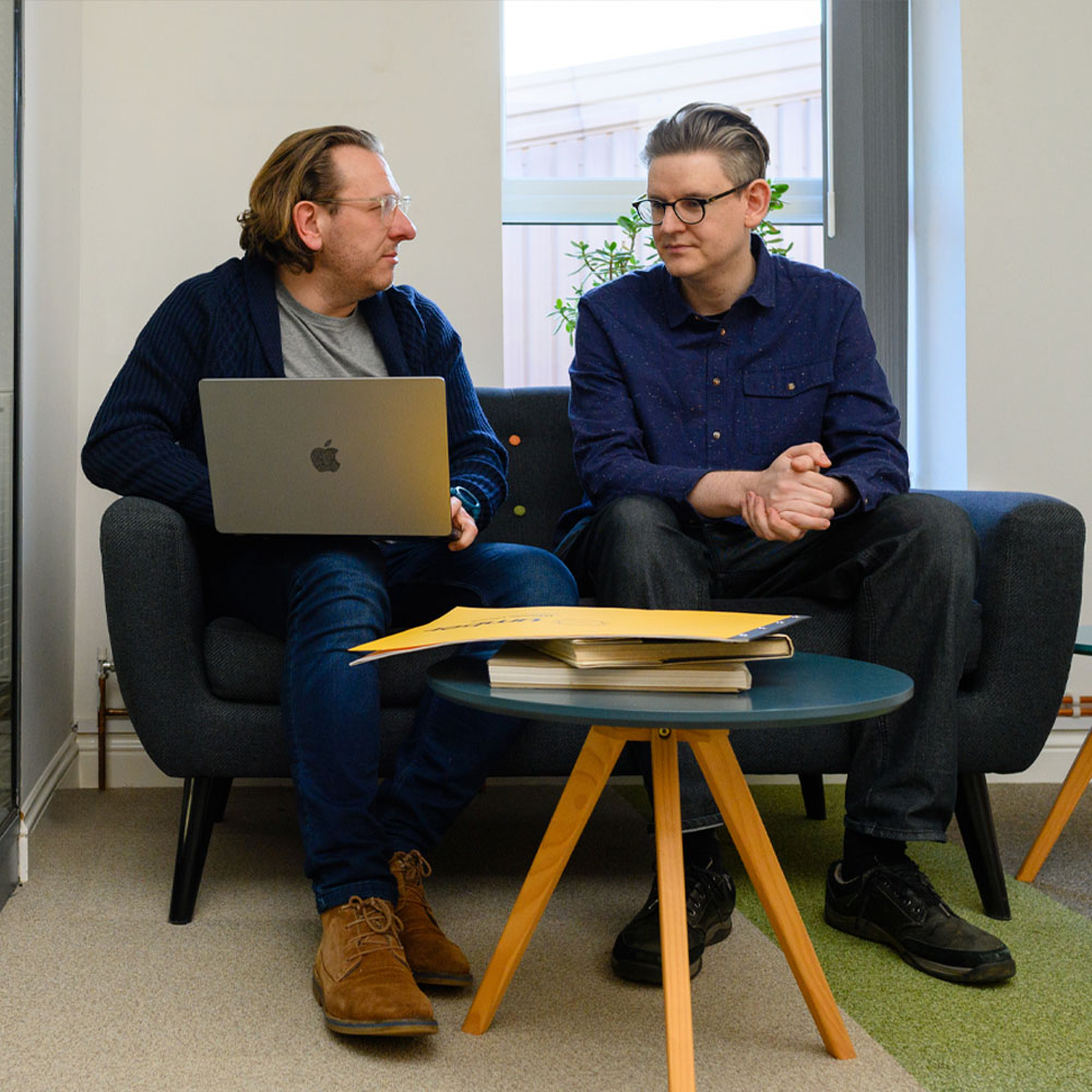 Two men sit on a sofa engaged in discussion. One holds a laptop, the other listens intently. A coffee table with books is in front.