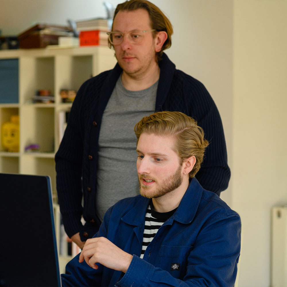Two men are focused on a computer screen in an office. One sits and types wearing a blue jacket, while the other stands behind, looking on.