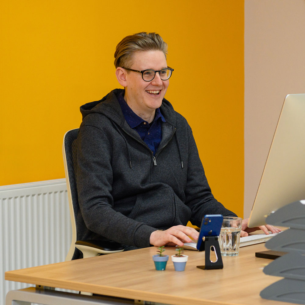 Smiling person with glasses, wearing a dark hoodie, seated at a desk with a computer, phone, and small plants, against a bright yellow wall.