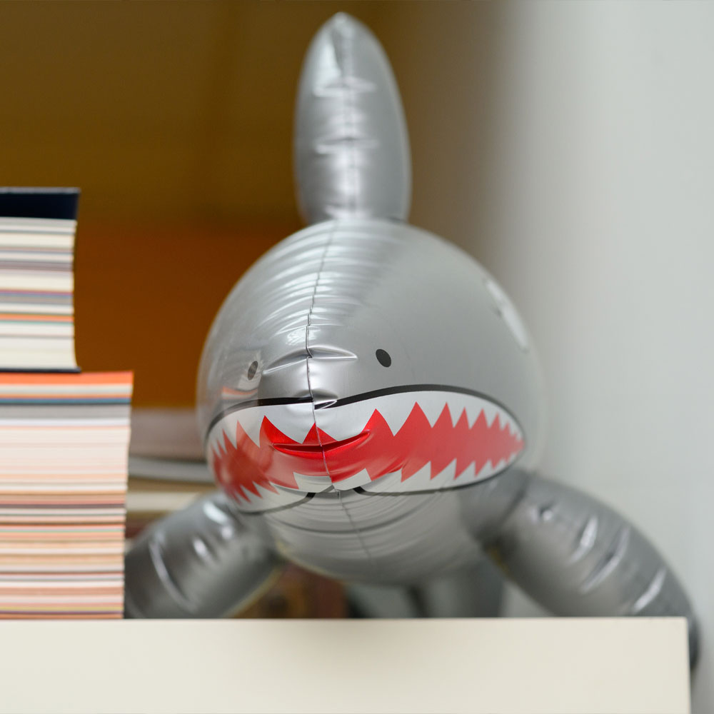 An inflatable grey shark toy with red zigzag mouth beside a stack of colourful magazines on a shelf.