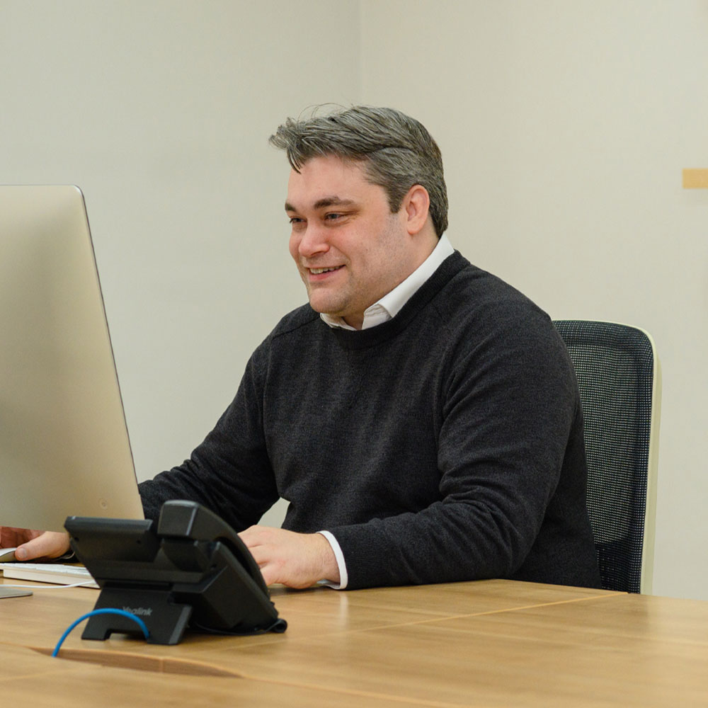Man smiling while working at a desk with a computer and phone in an office setting. He wears a dark sweater, creating a professional, positive mood.