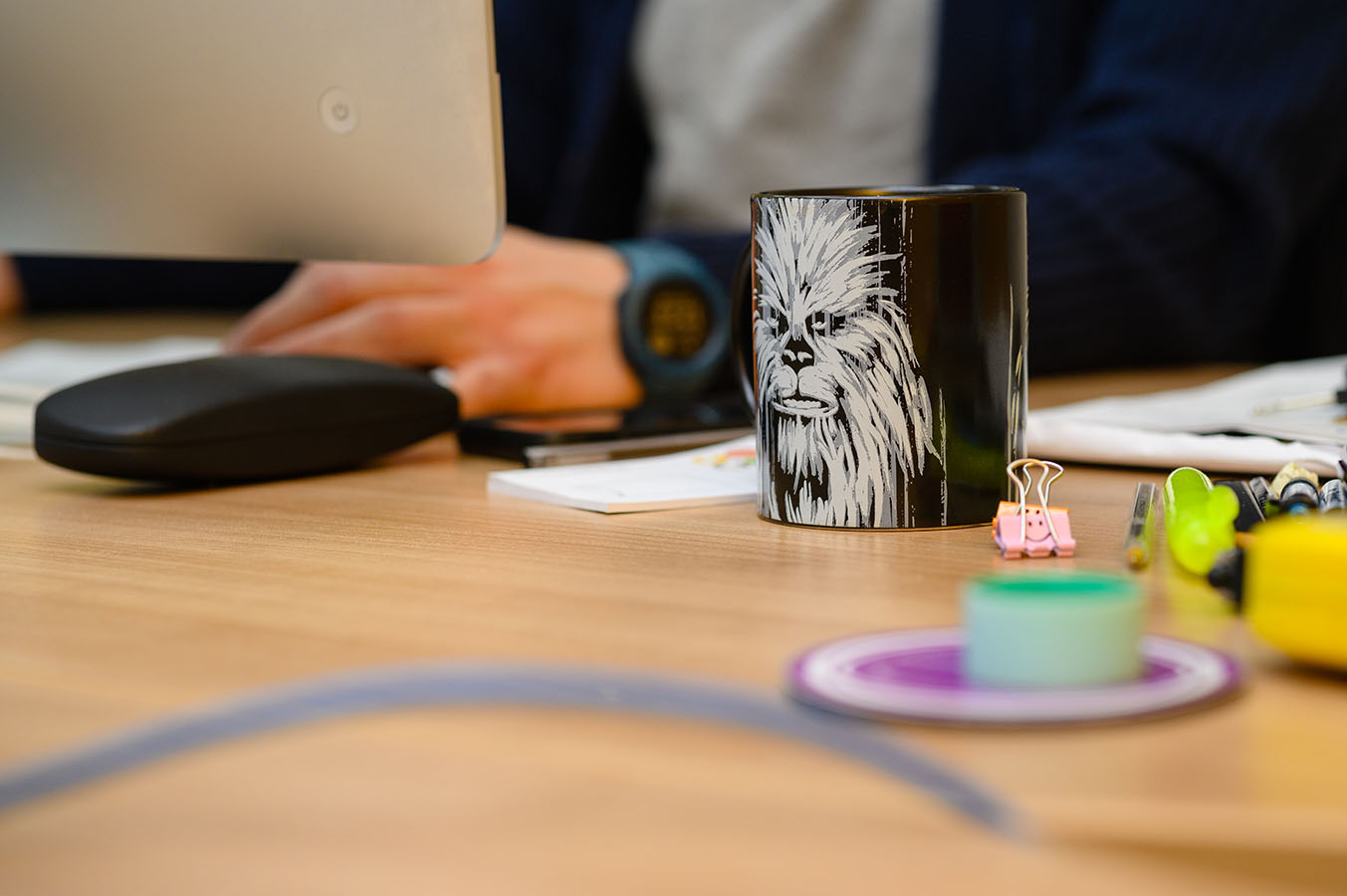 A person working at a desk with a black mug featuring a graphic of Chewbacca. Nearby are colourful binder clips and stationery, creating a casual office vibe.