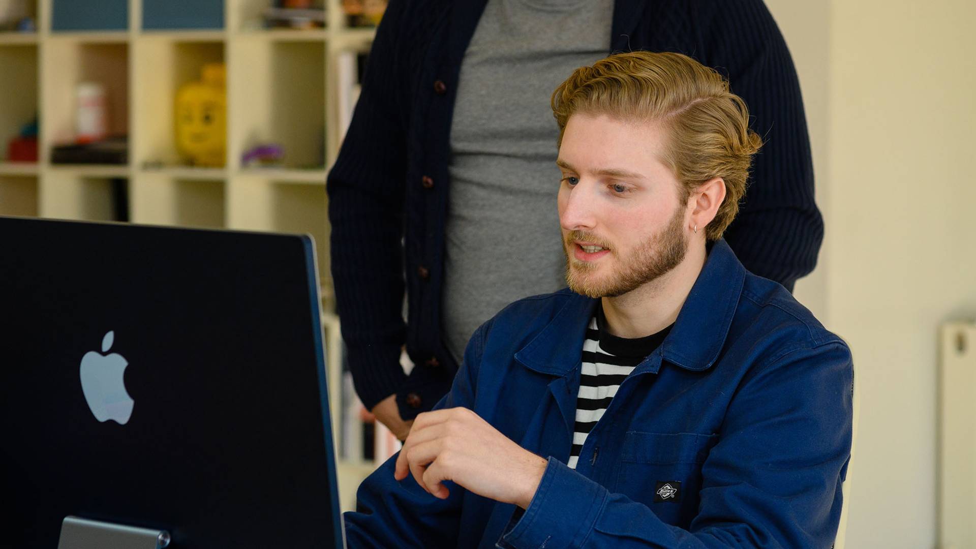A man with a beard sits focused at an Apple computer, wearing a blue jacket and striped shirt. Another person stands nearby, suggesting a collaborative work setting.