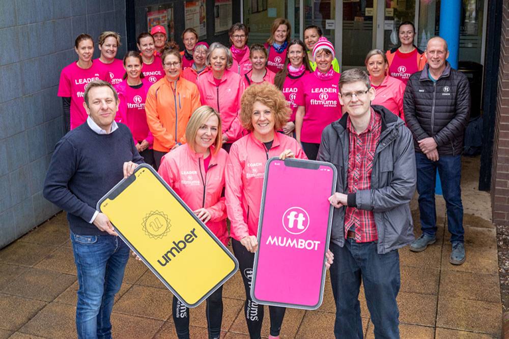 A group of smiling people, mostly women, wearing pink 'Fitmums' shirts, stand outside a building. Two people at the front hold large phone-shaped signs for apps 'Umber' and 'Mumbot.'