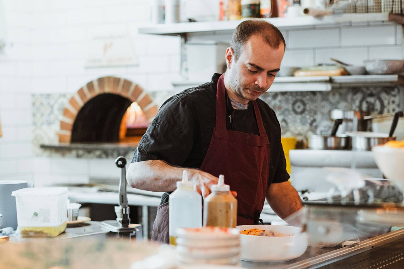 Chef in a maroon apron focuses on preparing a dish in a cozy, modern kitchen with tiled walls and a brick oven, conveying concentration and craftsmanship.