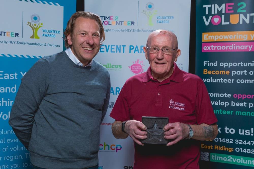 Two men smiling in front of 'Volunteer Awards' banners. The man on the left is in a grey sweater; the man on the right in a red shirt holds a star-shaped award.