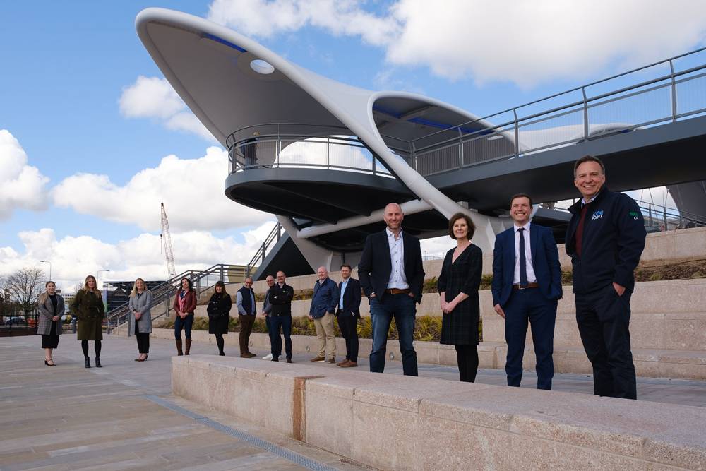 A group of people stand in front of a modern, curved structure under a blue sky with clouds. They appear cheerful and relaxed, suggesting teamwork or collaboration.