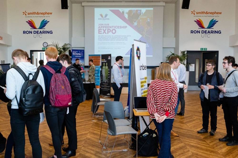 People mingling at an apprenticeship expo in a hall with banners and displays. The atmosphere is vibrant and interactive, fostering networking.