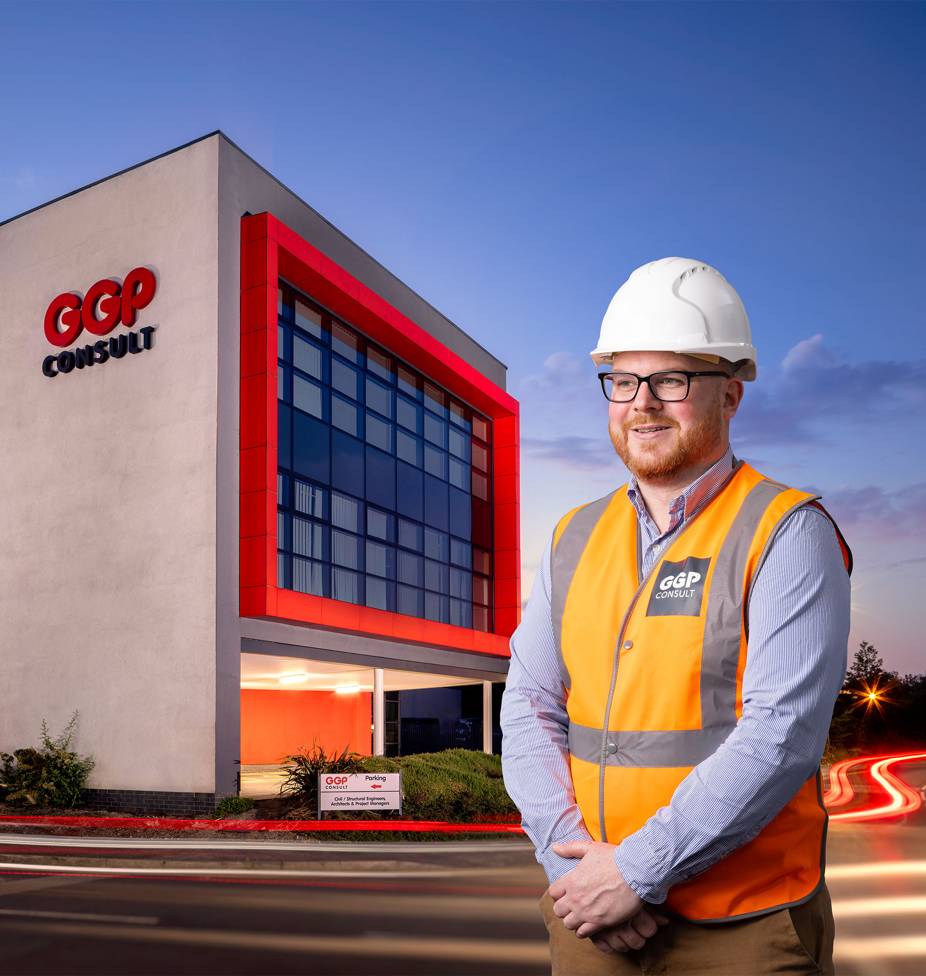 Smiling man in a hard hat and orange vest stands in front of a modern building with a red-framed facade at dusk. The building is labeled "GGP Consult."