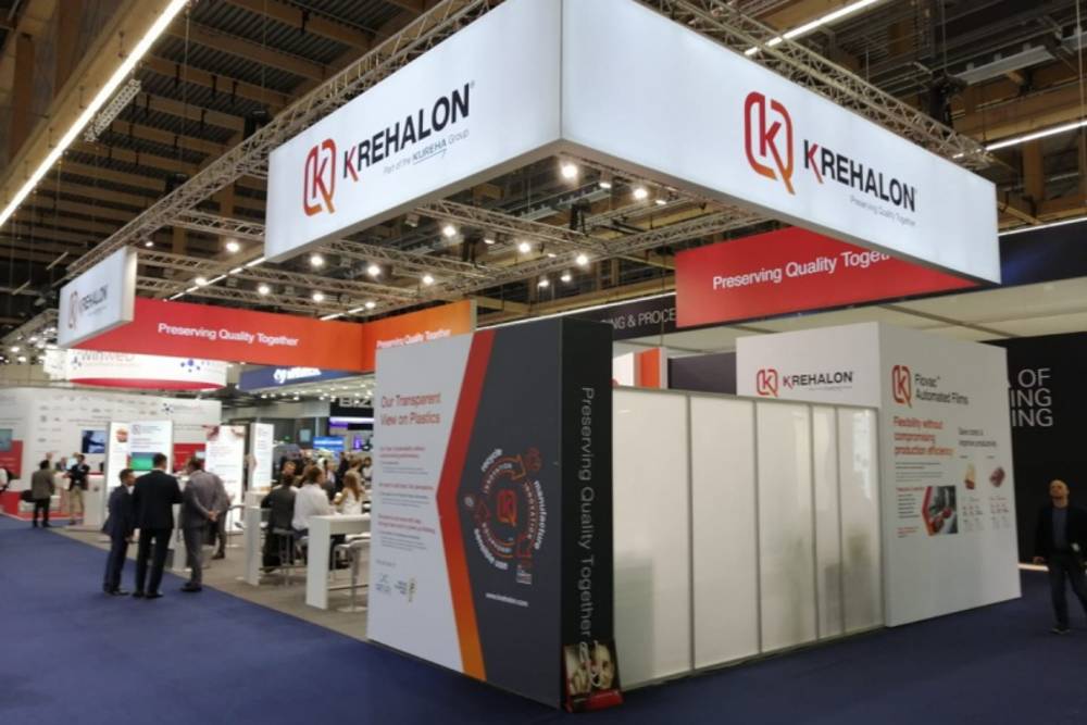 Trade show booth for Krehalon with bright signs, including 'Preserving Quality Together.' People engaging and reading displays, creating a professional atmosphere.
