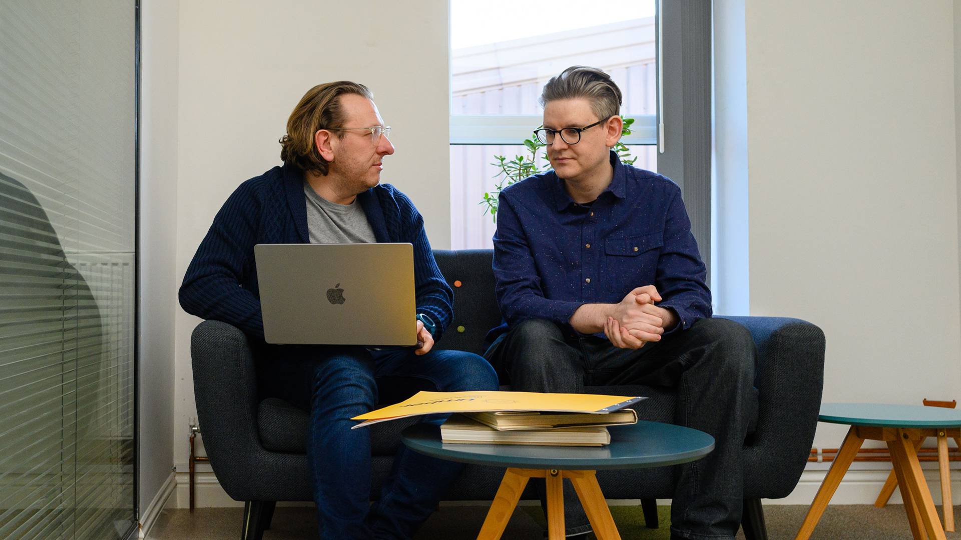Two men sit on a couch in a bright office, engaged in conversation. One holds a laptop, and papers are on a table.