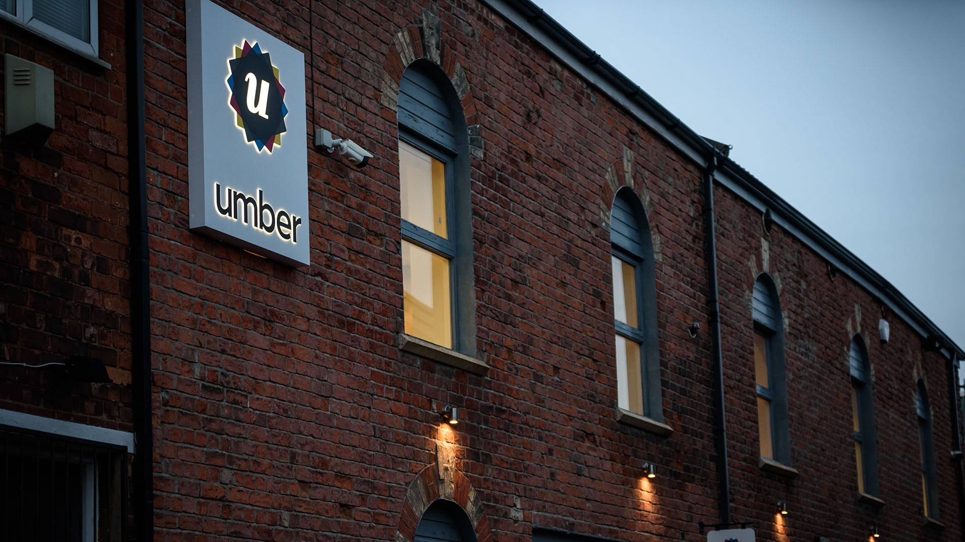 Red brick building with tall arched windows glowing warmly. A sign with 'umber' and a 'u' logo is illuminated, giving an inviting, cozy vibe.