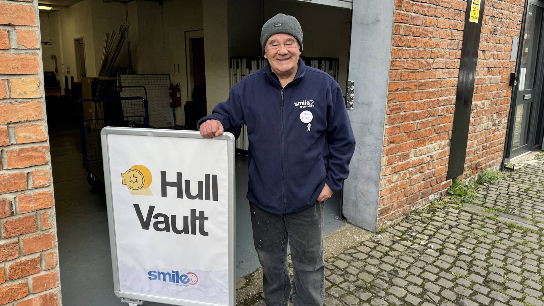 A smiling person in a beanie and jacket stands next to a sign for 'Hull Vault' by a brick building.