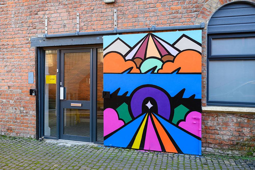 Colorful geometric mural on brick wall beside a glass door and window. Features mountains, sunbursts, clouds, and a vibrant rainbow road.