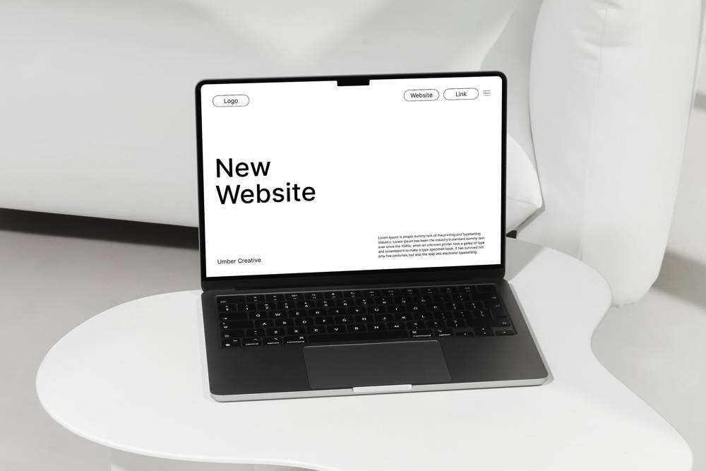 A sleek laptop on a white table displays a minimalistic webpage with "New Website" in bold text. The setting is modern and clean.