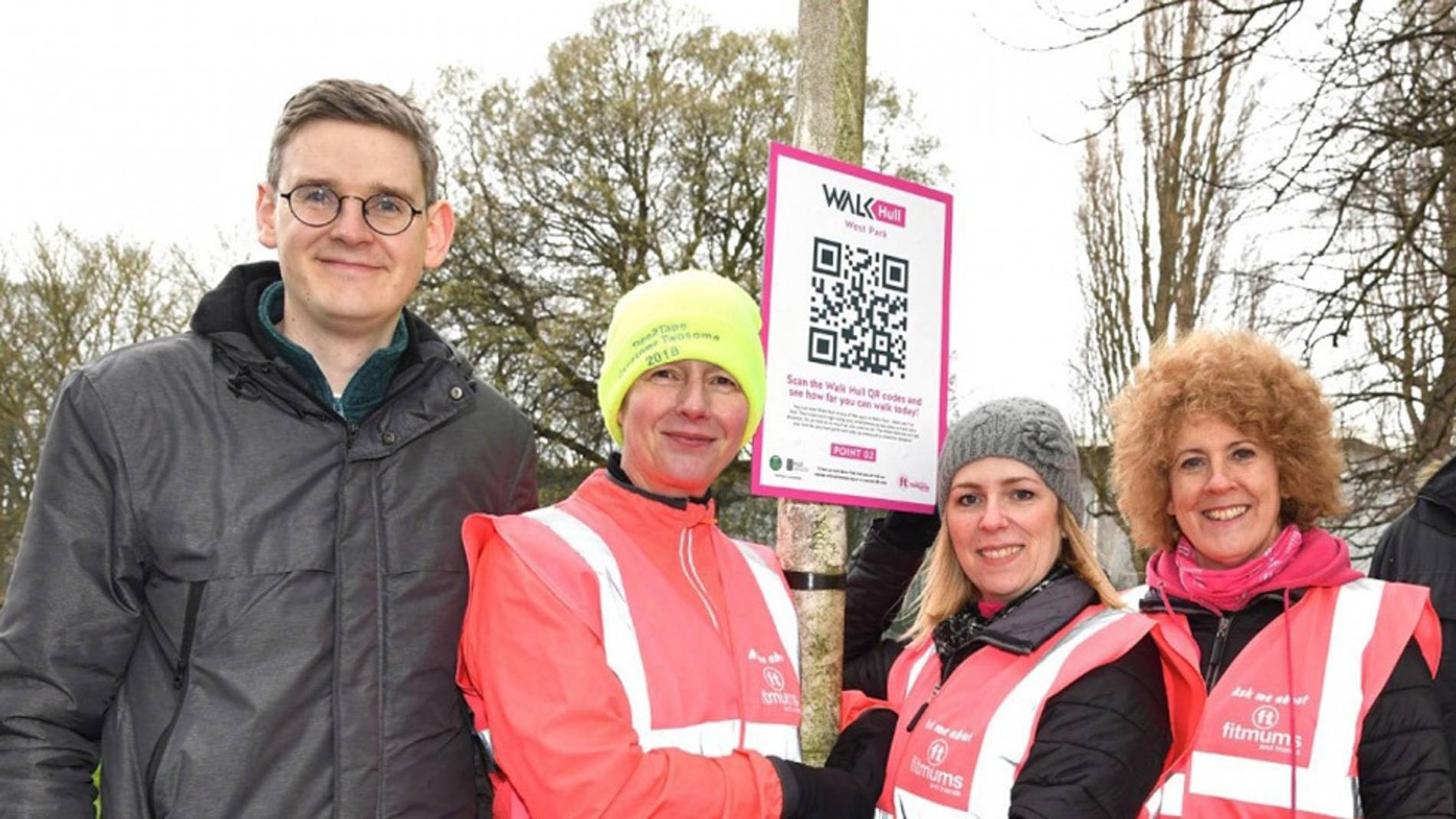 A group of four smiling adults pose outdoors in winter clothing. Three wear pink safety vests; one holds a 'Walk Hull' QR code sign.
