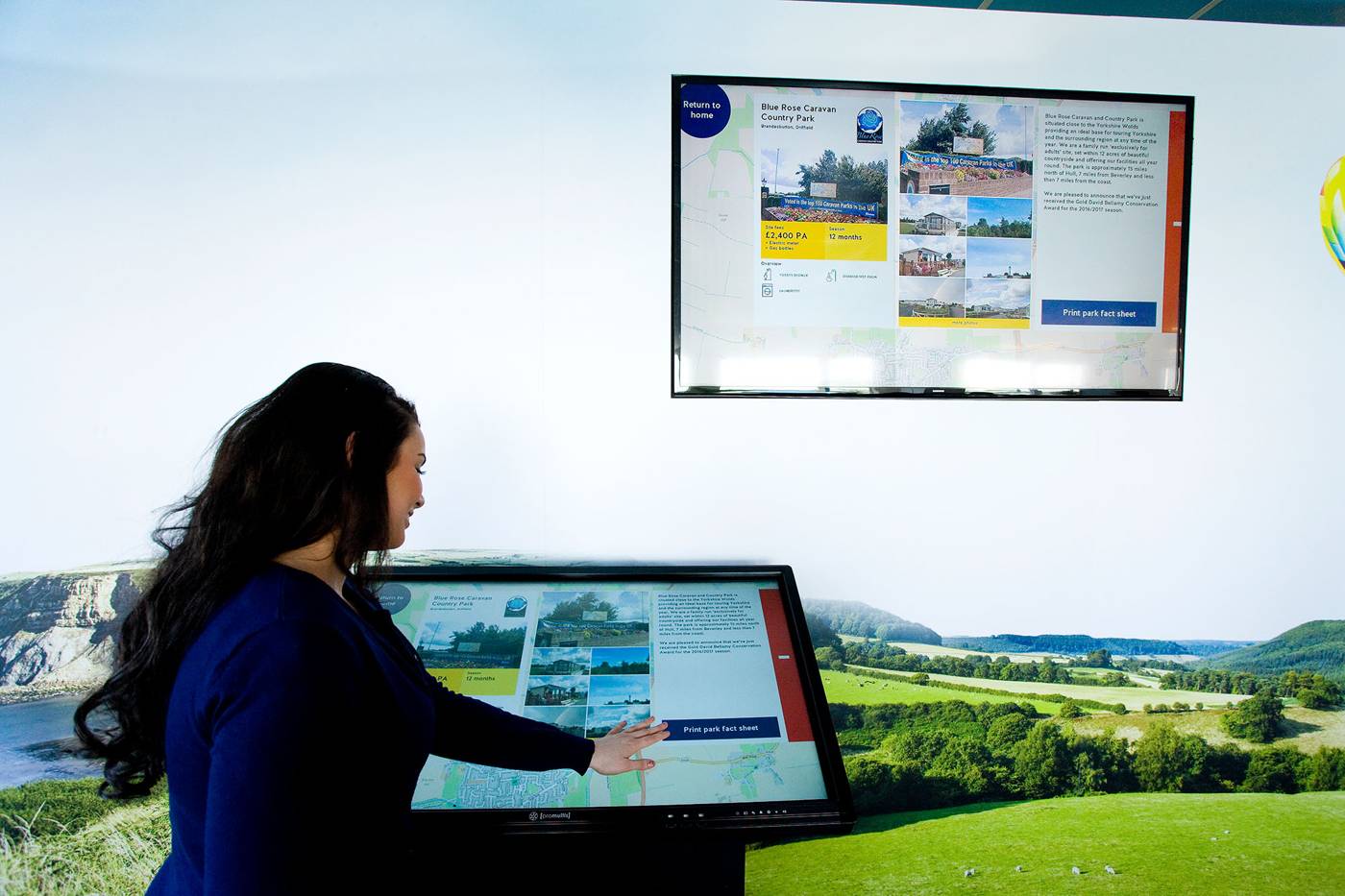 A woman in a blue shirt interacts with a digital touchscreen displaying scenic park information. In the background, a screen shows similar content, set against a picturesque landscape of rolling green hills, conveying a sense of exploration and technology