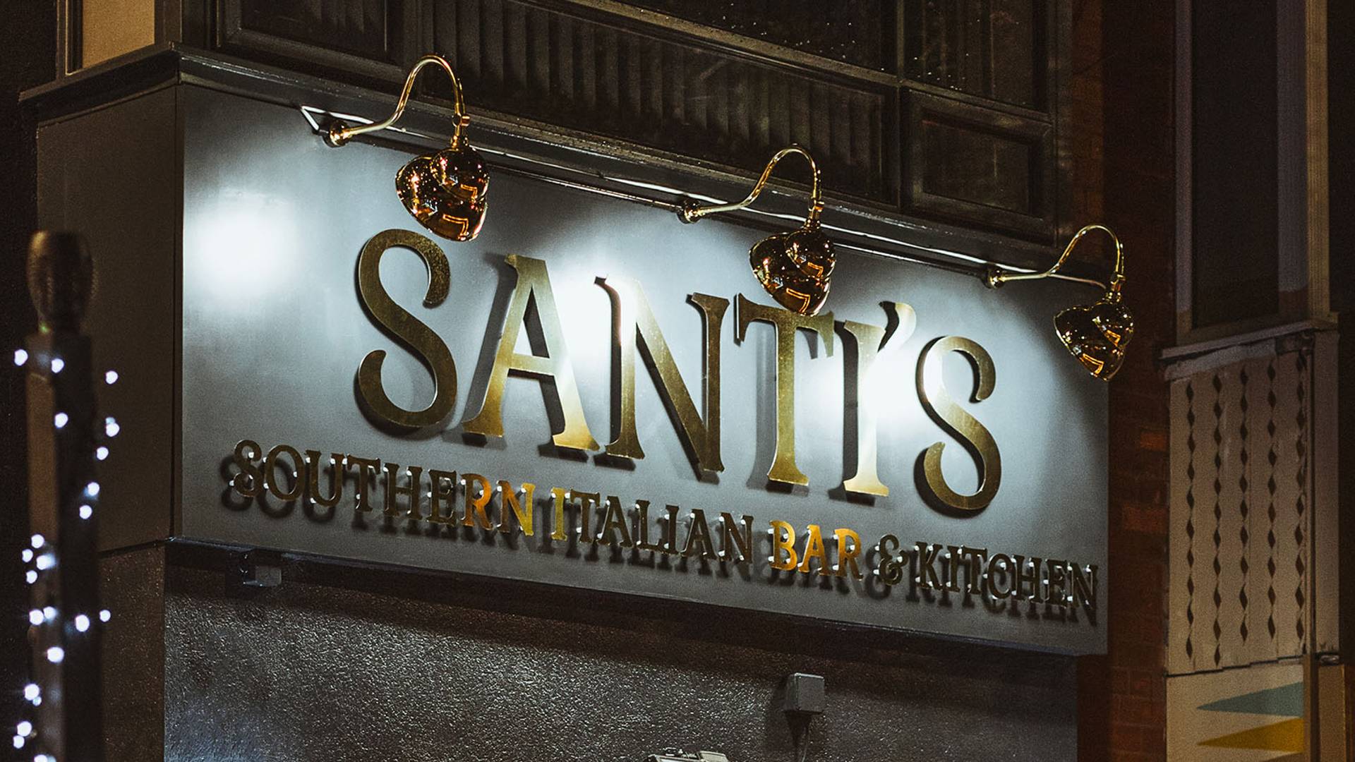 Sunny day view of a brick building with a decorative lowercase 'b' mounted on its wall. Sunlight and tree foliage are visible against a clear blue sky.