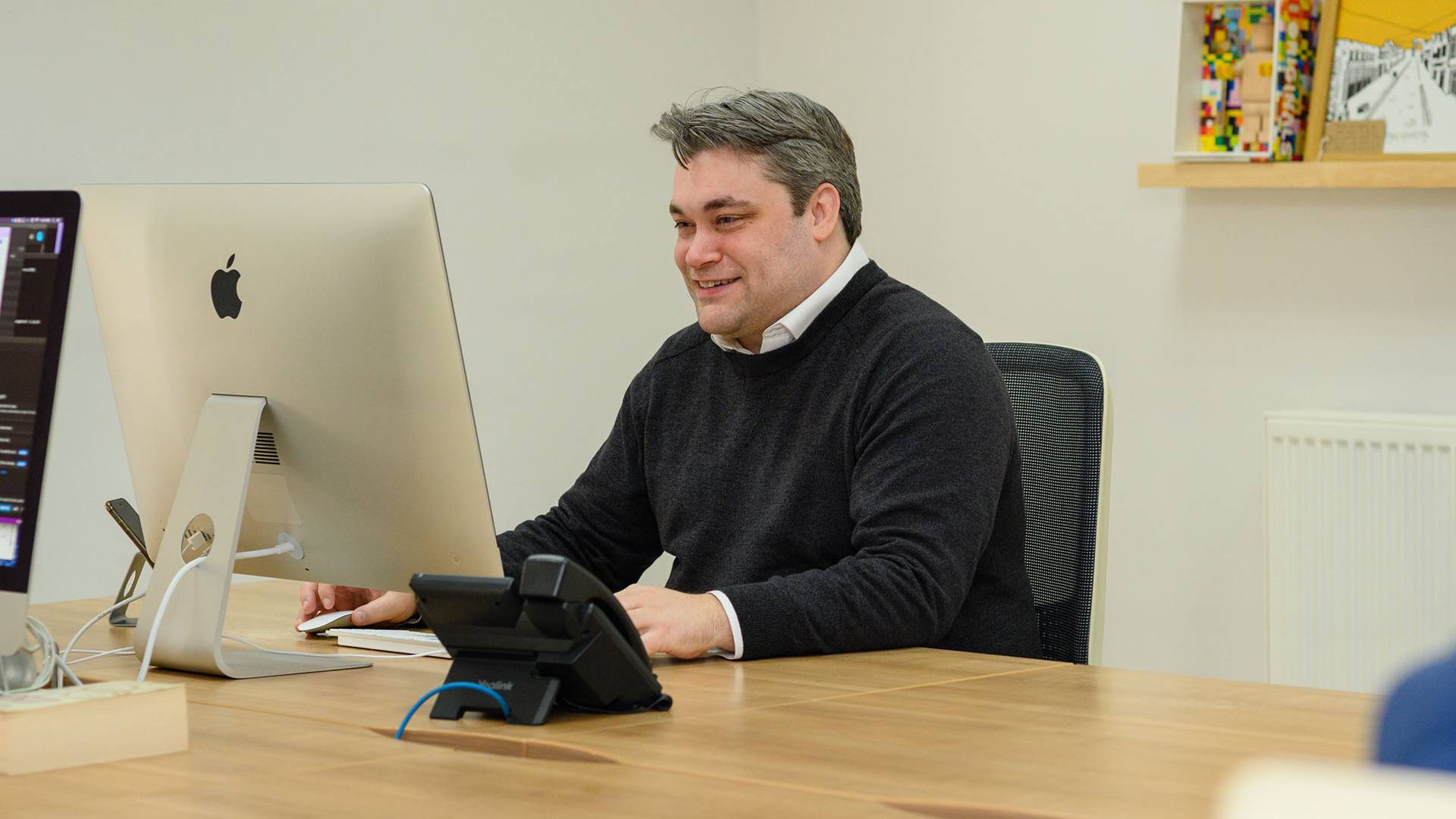 Smiling man sitting at a wooden desk working on a large computer. Office setting with a phone dock and shelf.