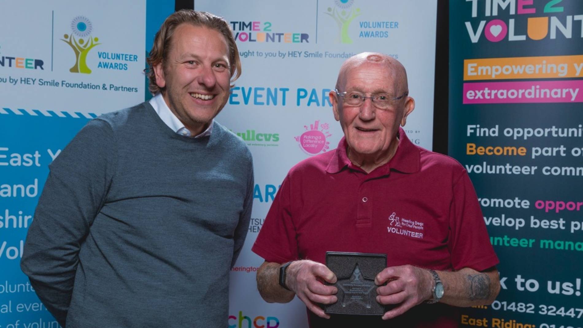 Two men smiling in front of 'Volunteer Awards' banners. The man on the left is in a grey sweater; the man on the right in a red shirt holds a star-shaped award.