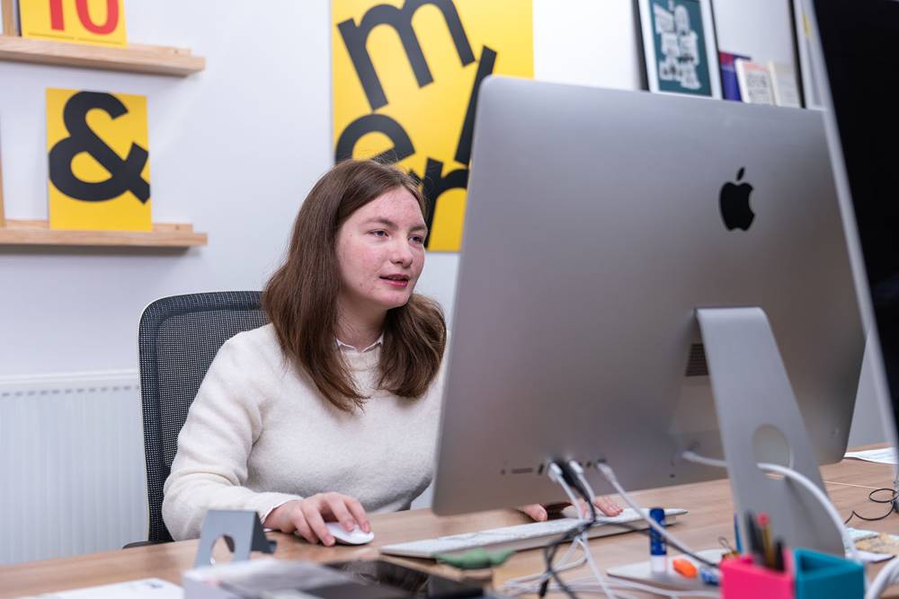 A woman in a white sweater works at a large desktop computer in a modern office. Yellow and black wall art is visible.
