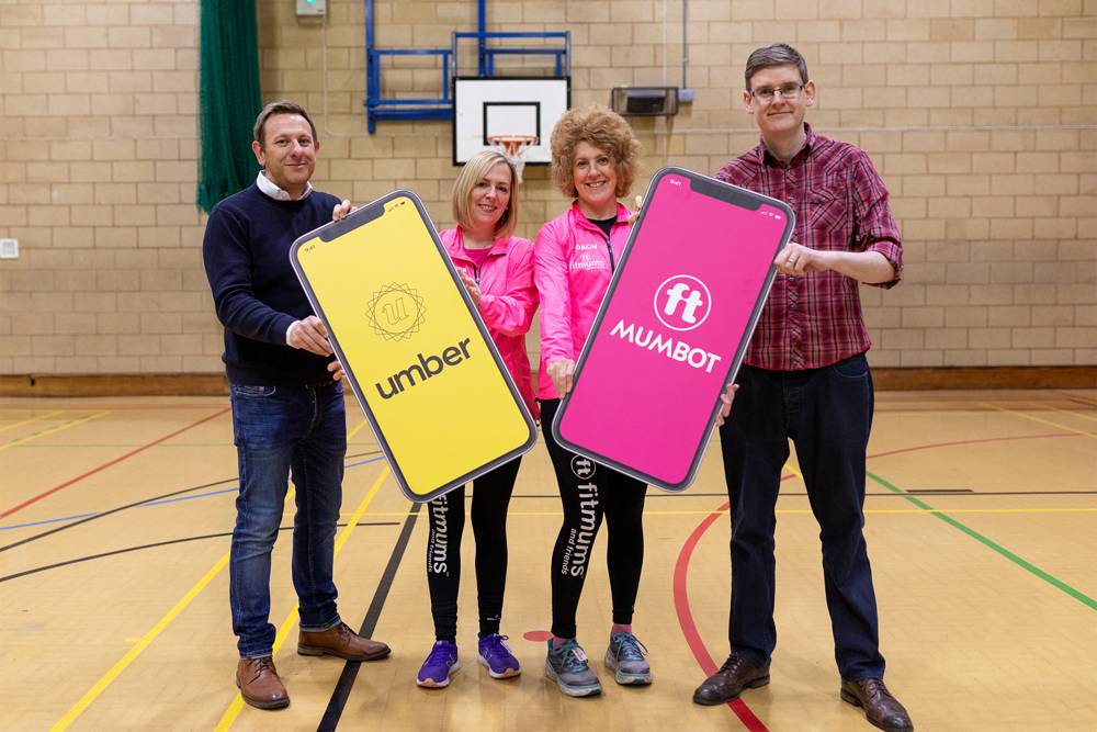 Four people stand in a gym, smiling and holding large smartphone-shaped signs. Two women wear bright pink jackets.