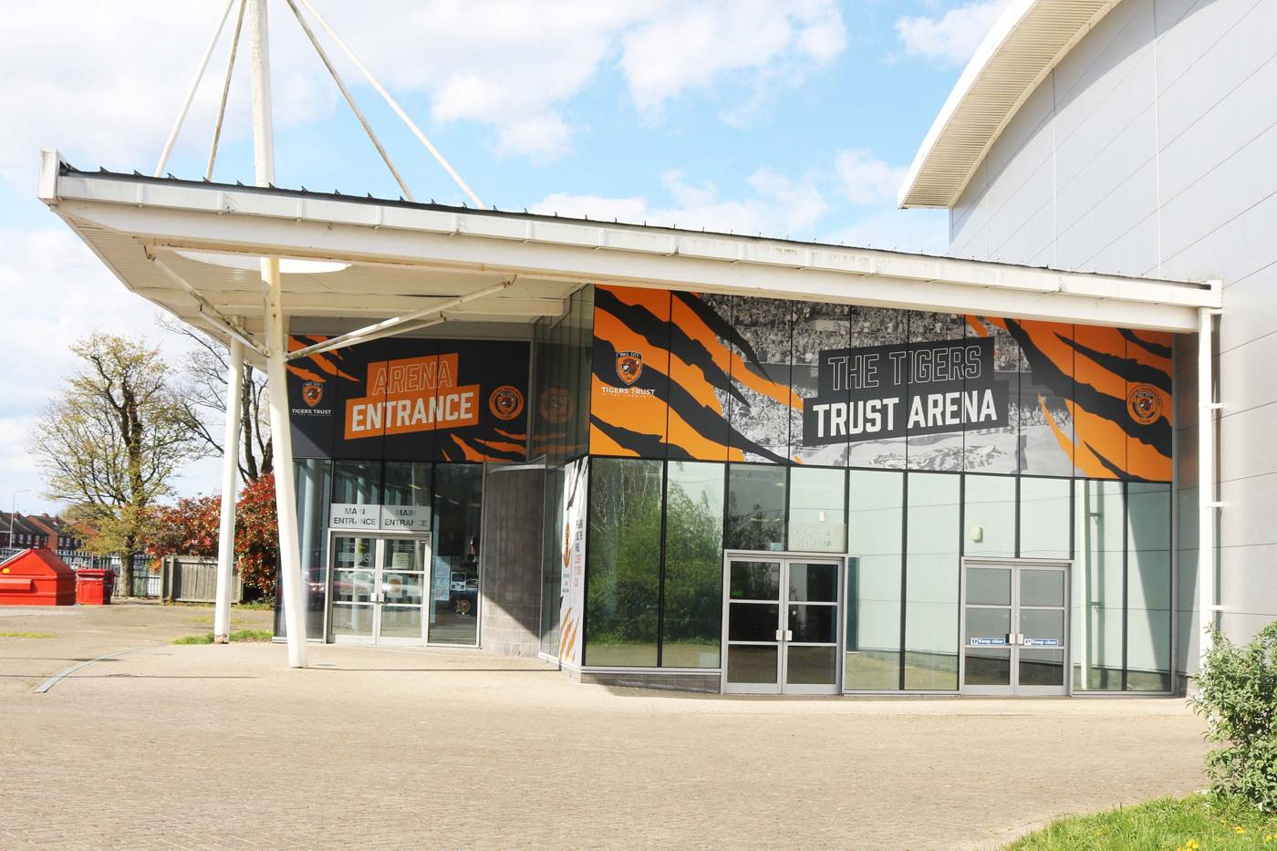 Entrance to The Tigers Trust Arena with bold orange and black tiger stripe design, featuring large glass doors and a clear sky backdrop.