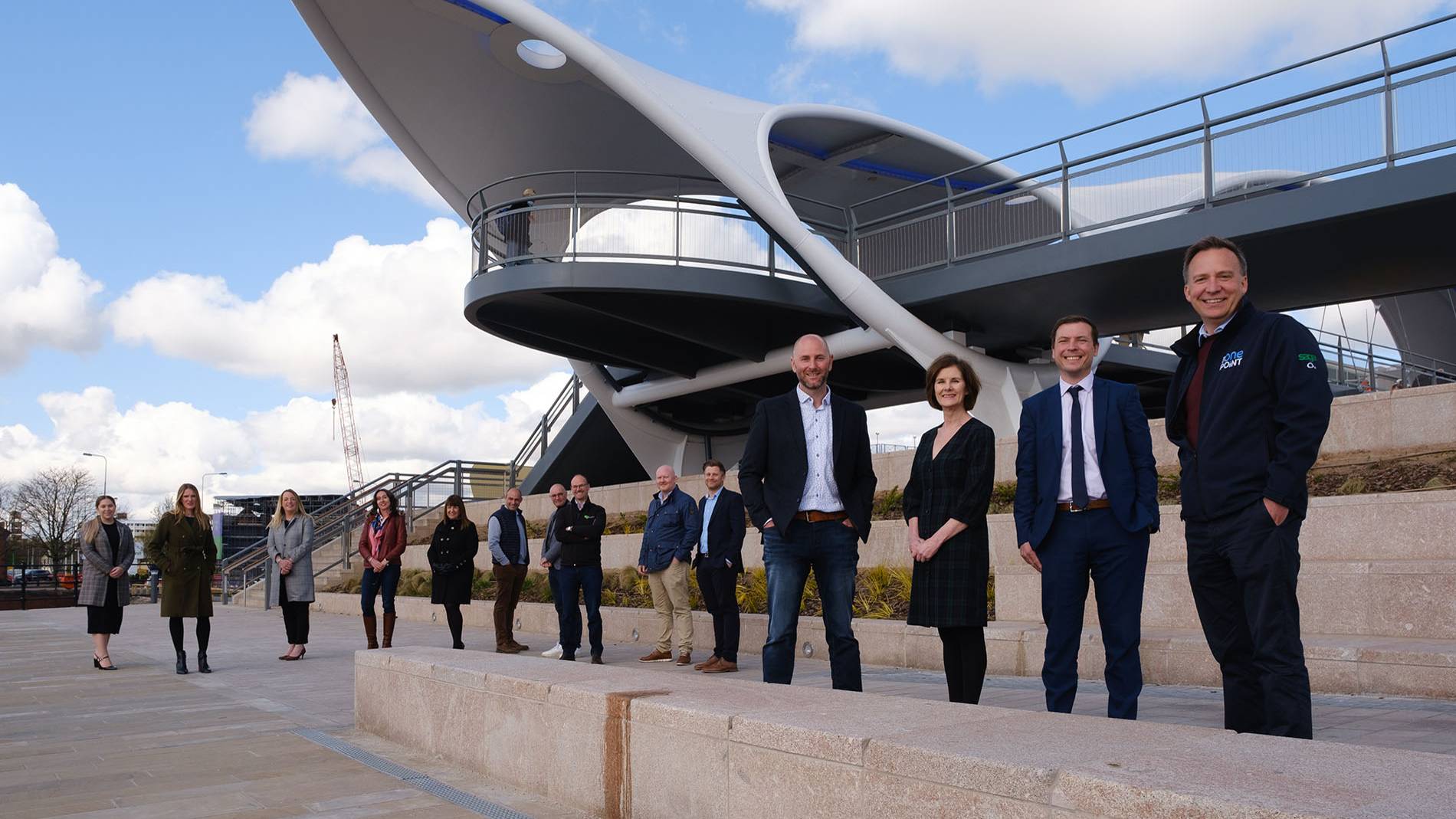 A group of people stand in front of a modern, curved structure under a blue sky with clouds. They appear cheerful and relaxed, suggesting teamwork or collaboration.