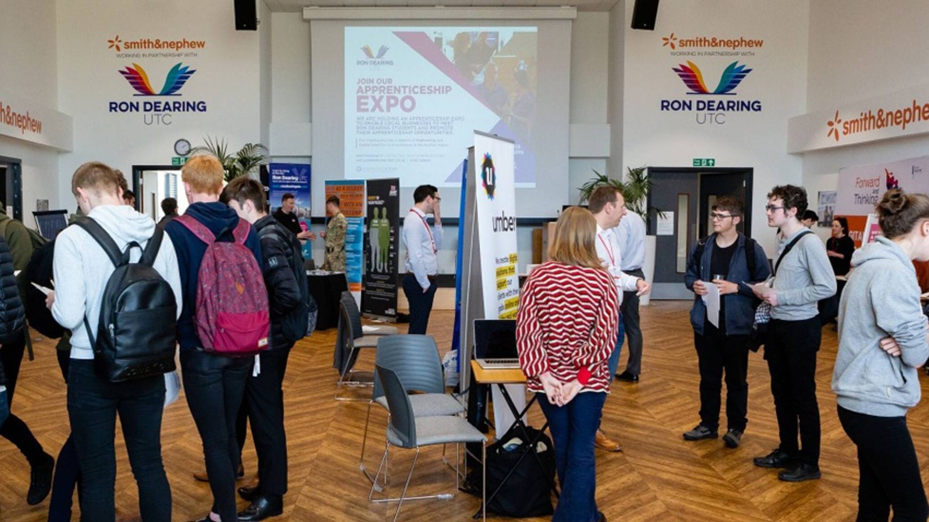 People mingling at an apprenticeship expo in a hall with banners and displays. The atmosphere is vibrant and interactive, fostering networking.