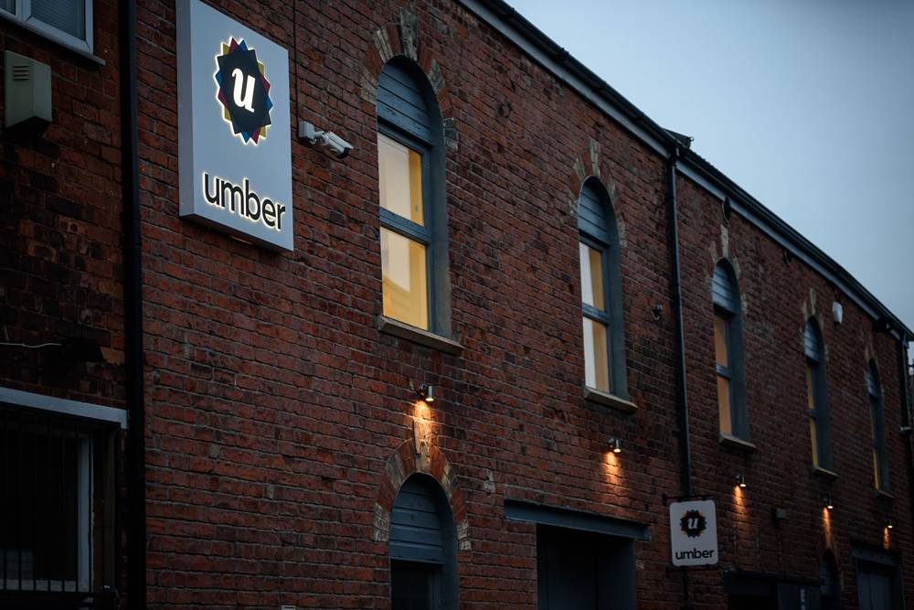 Red brick building with tall arched windows glowing warmly. A sign with 'umber' and a 'u' logo is illuminated, giving an inviting, cozy vibe.
