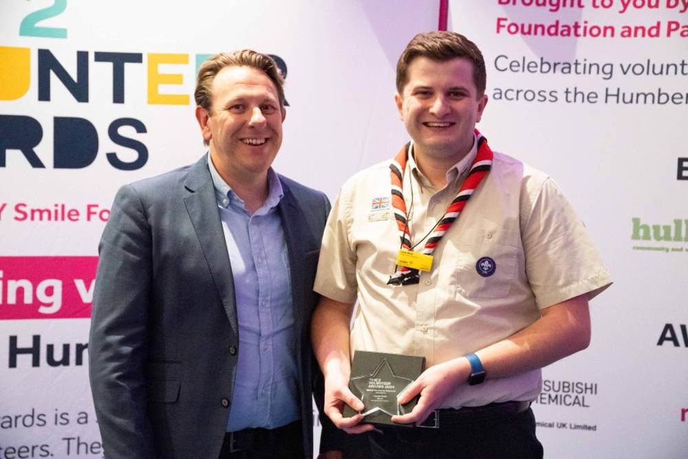Two men smile at an awards event. One holds an award plaque and wears a scout uniform. Background shows colorful event banners and text.