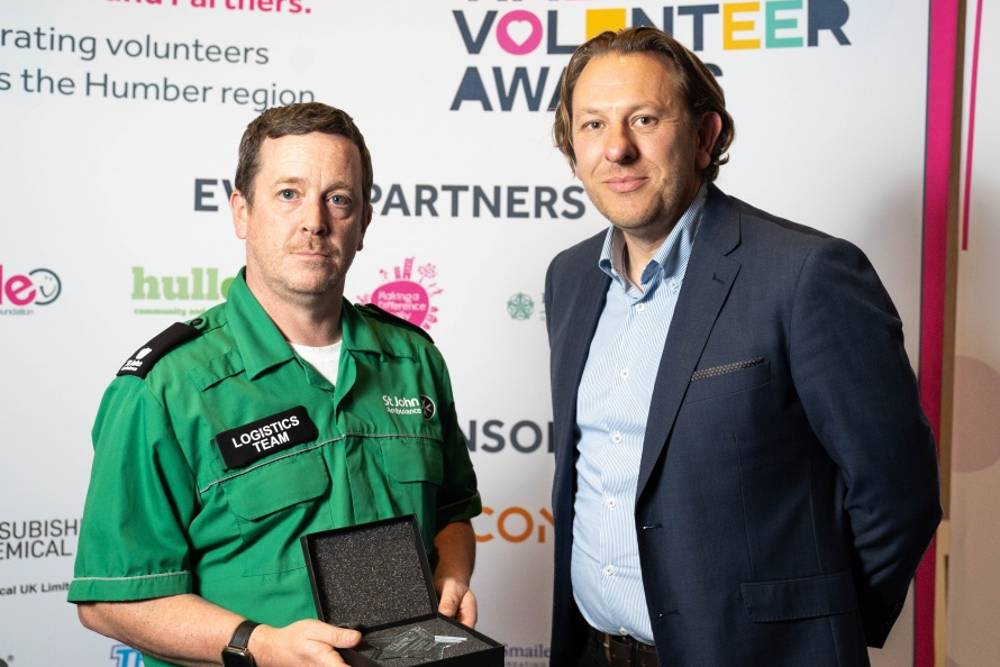 Two men at a volunteer awards event. One in a green logistics team uniform holds a plaque. Background features event logos and the text 'Volunteer Awards'