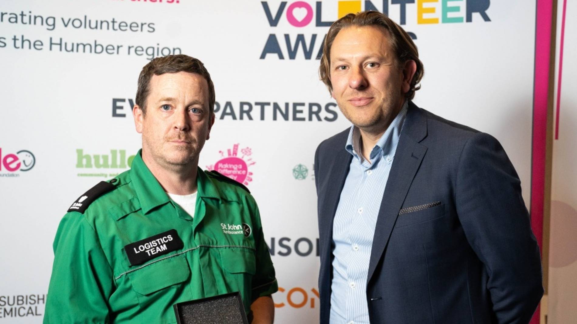 Two men at a volunteer awards event. One in a green logistics team uniform holds a plaque. Background features event logos and the text 'Volunteer Awards'