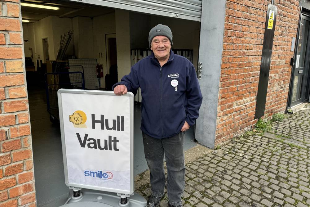 A smiling person in a beanie and jacket stands next to a sign for 'Hull Vault' by a brick building.