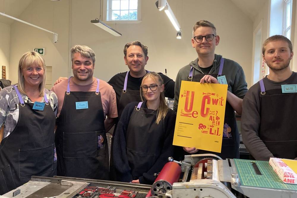 A group of six people wearing black aprons stands in a well-lit workshop. They are smiling, and one person holds a bright yellow art print.